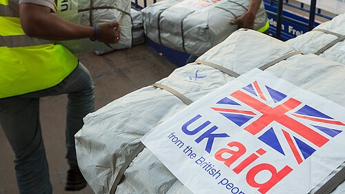 Two men are moving parcels wrapped in white plastic. In the foreground, a label on one package is visible reading "UK Aid from the British people" and a union flag. Image - Dfid CCBY-ND2.0