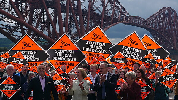Scottish Liberal Democrat leader Alex Cole-Hamilton. Behind him is a crowd holding Scottish Liberal Democrat diamond signs. In the background is the Forth rail bridge.