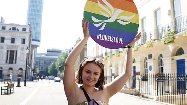 Young person holding a round sign over their head with a rainbow-striped, pride flag design overlaid with the Liberal Democrat bird of liberty emblem and #LoveIsLove