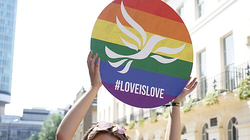 Young person holding a round sign over their head with a rainbow-striped, pride flag design overlaid with the Liberal Democrat bird of liberty emblem and #LoveIsLove