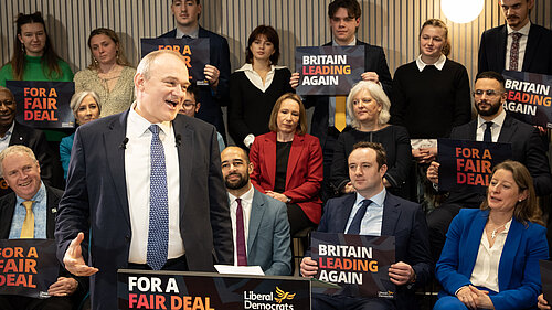 Ed Davey making a speech, a crowd of people are in the background holding signs reading "Britain Leading Again" and "For a Fair Deal"