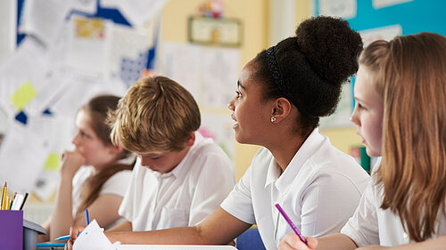 Four pupils sat at a table in school, looking at their books or up at out of shot.