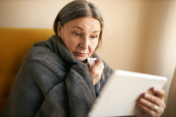 Older woman wrapped in a blanket. She is holding a tissue and looking at an electronic tablet. She looks concerned.