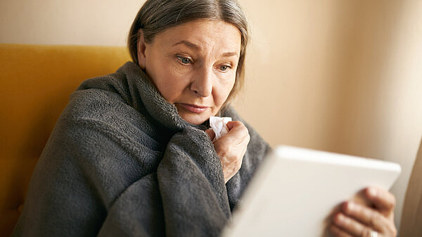 Older woman wrapped in a blanket. She is holding a tissue and looking at an electronic tablet. She looks concerned.