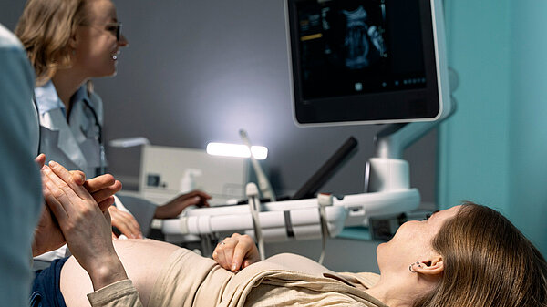 In the foreground woman is lying down in a medical setting, looking up at a screen showing an ultrasound. On the left of the photo, is a nurse operating the ultrasound.