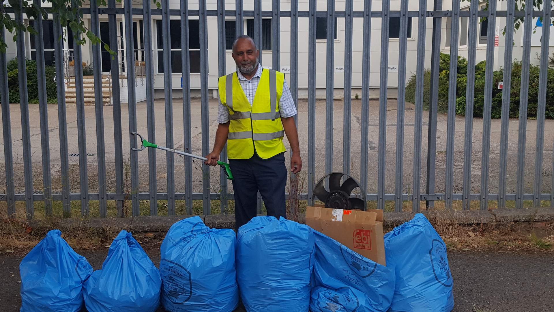 Cllr Mo Fayyaz wearing a high vis vest and holding a litter picker. In front of him is a row of blue bin bags with rubbish collected on the litter pick.